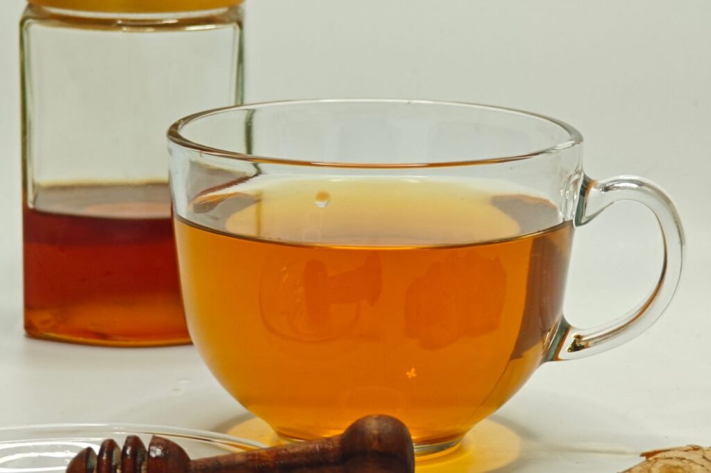 Glass cup of amber ginger tea next to a jar of honey, a honey dipper and a knob of fresh ginger root