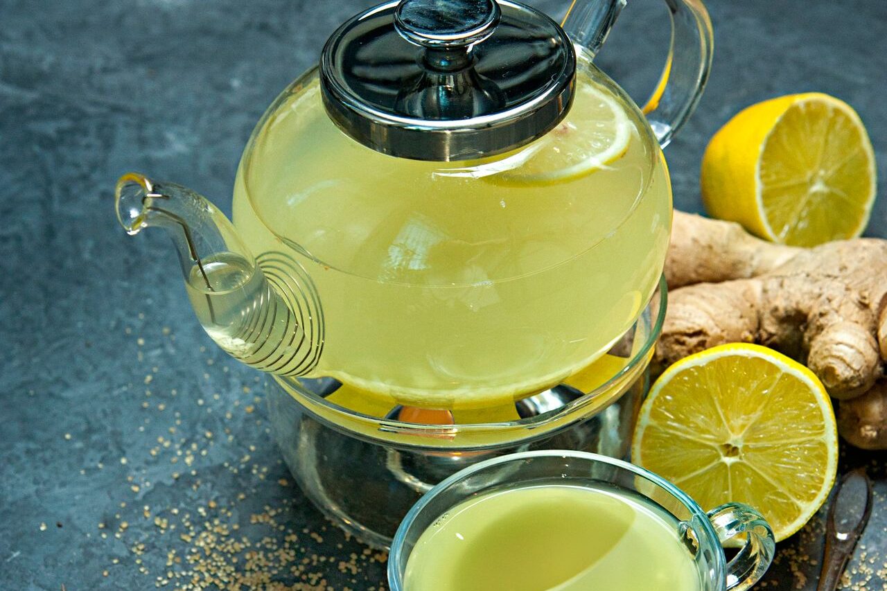 Glass teapot and cup of fresh lemon ginger tea with lemon halves and ginger root on a dark table