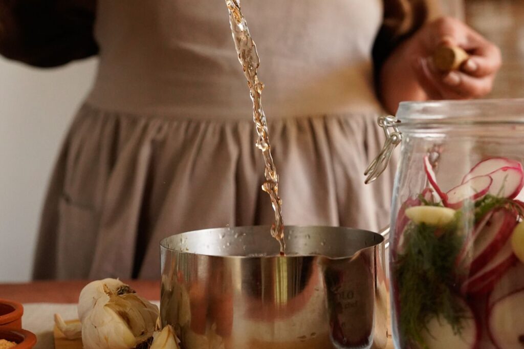 Person in an apron pouring apple cider vinegar from a glass bottle into a jar of chopped ingredients for fire cider
