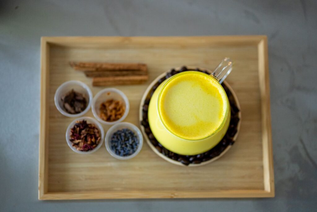 Top-down view of a frothy golden milk latte in a yellow cup on a wooden tray with small dishes of cinnamon, cardamom and other spices