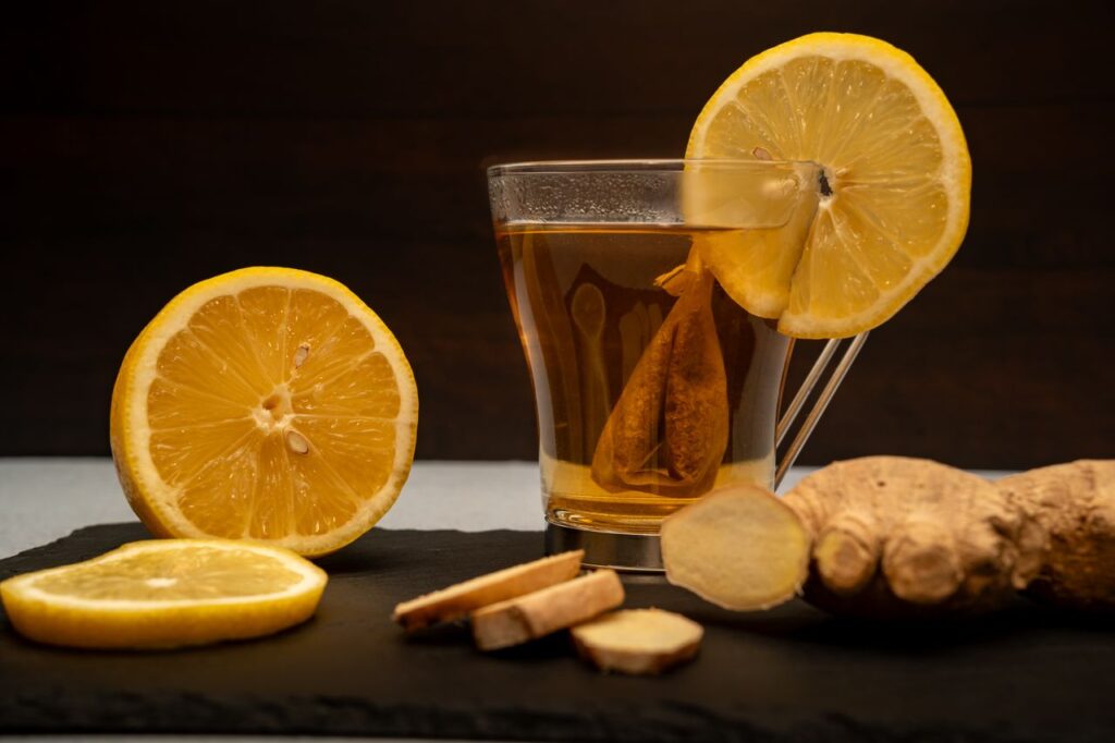 Glass of hot lemon ginger tea with a lemon slice on the rim, surrounded by halved lemons and fresh ginger root on dark slate