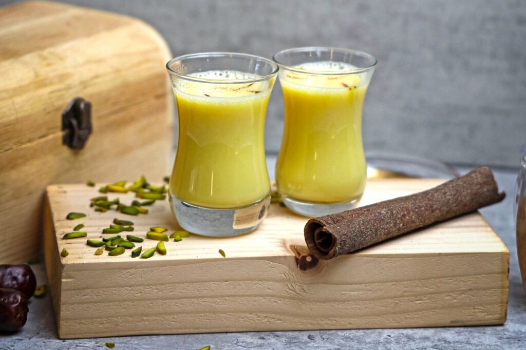 Two glasses of golden turmeric milk on a wooden board with a cinnamon stick and chopped pistachios