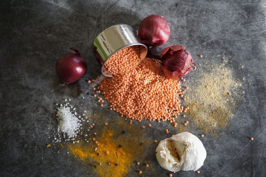 Red lentils spilling from a tin beside red onions, garlic and spices