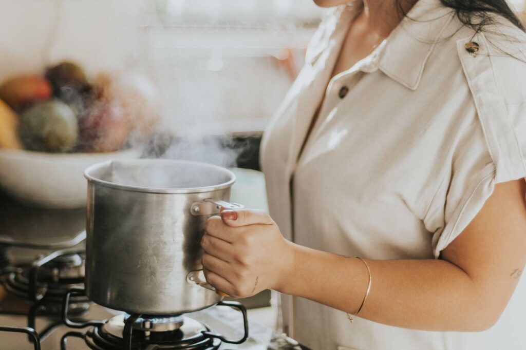 Person heating a stainless steel pot of water on a gas hob in a home kitchen with steam rising
