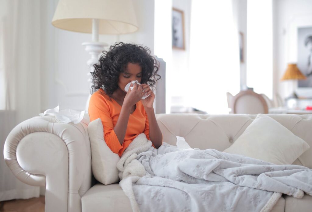 Woman wrapped in a grey blanket on a sofa blowing her nose into a tissue