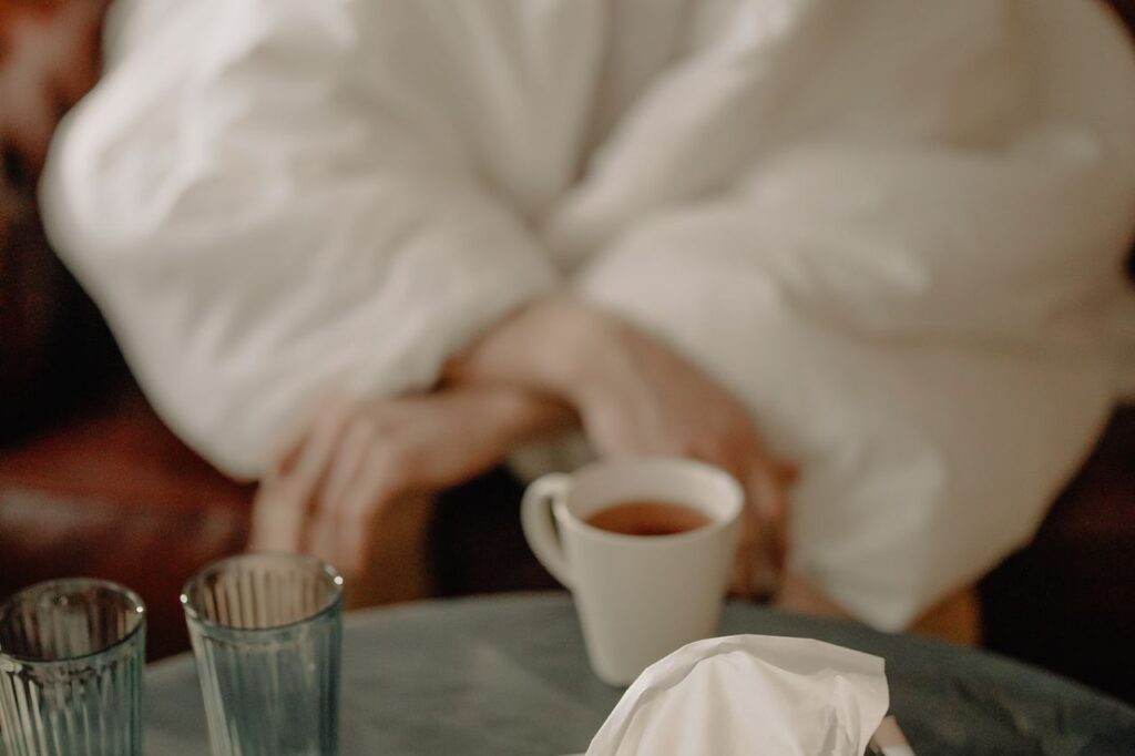 Person wrapped in a white duvet holding a mug of tea on a sofa with a box of tissues, resting with a cold
