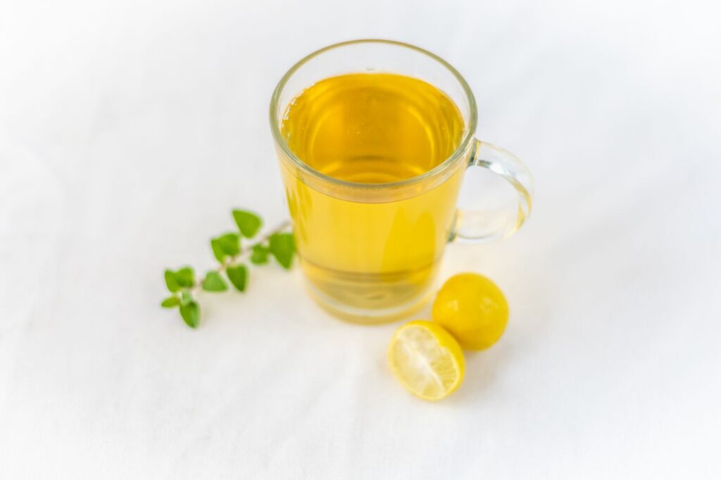 Clear glass mug of pale yellow herbal tea beside fresh herb sprigs and small lemons on a white surface
