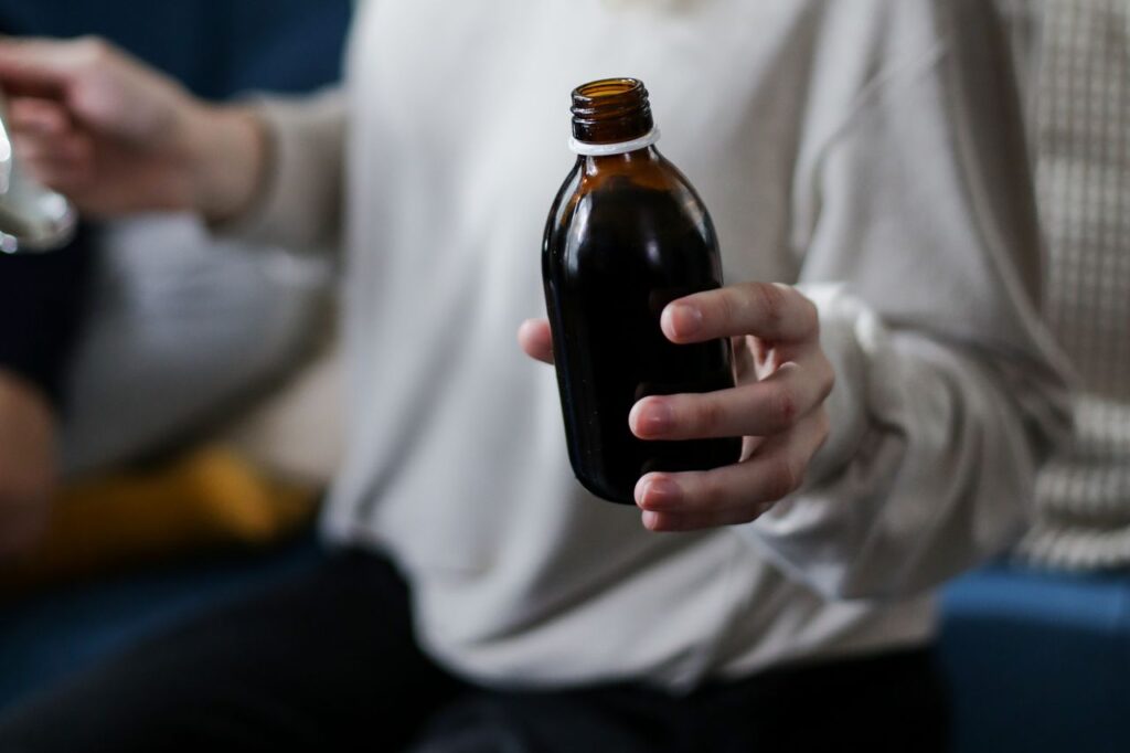 Person holding an open amber glass bottle of dark syrup, ready to measure out a daily dose