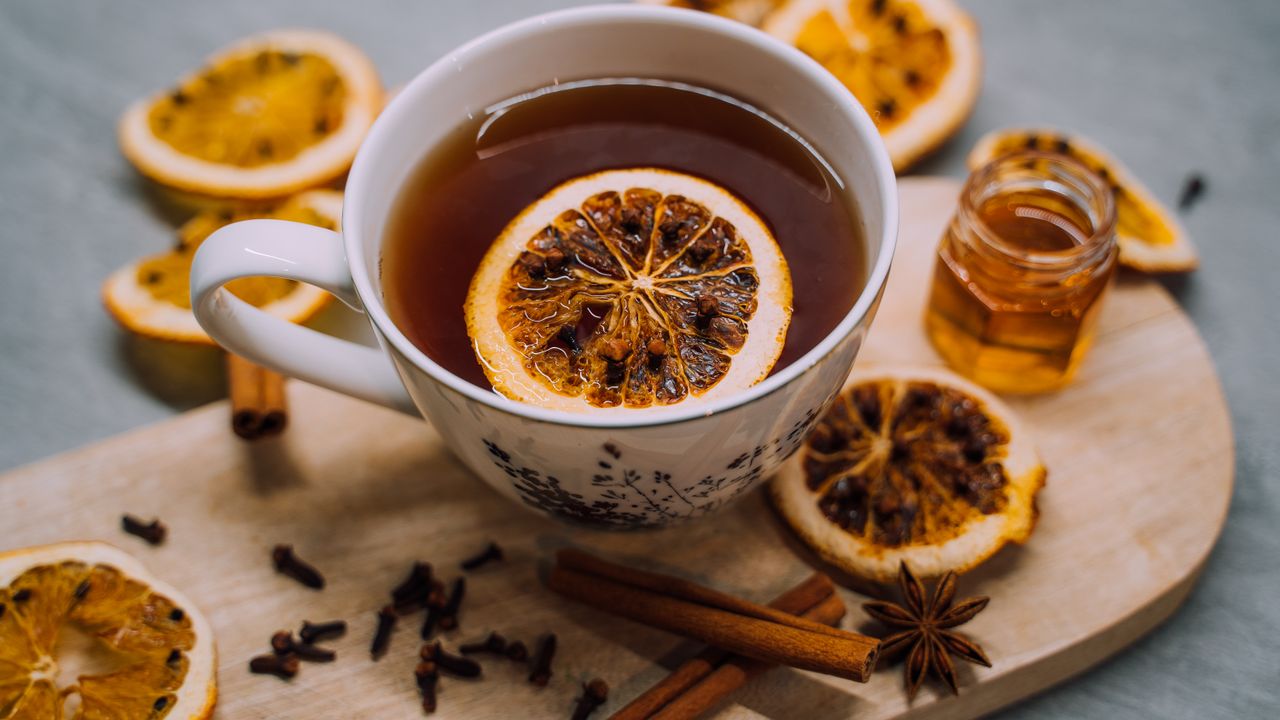 Cup of hot tea with a dried orange slice, cinnamon sticks, cloves and a jar of honey on a wooden board, a classic sore throat remedy
