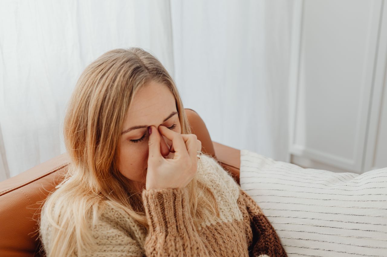 Woman on a sofa pinching the bridge of her nose in sinus pain, a common reason to try a neti pot