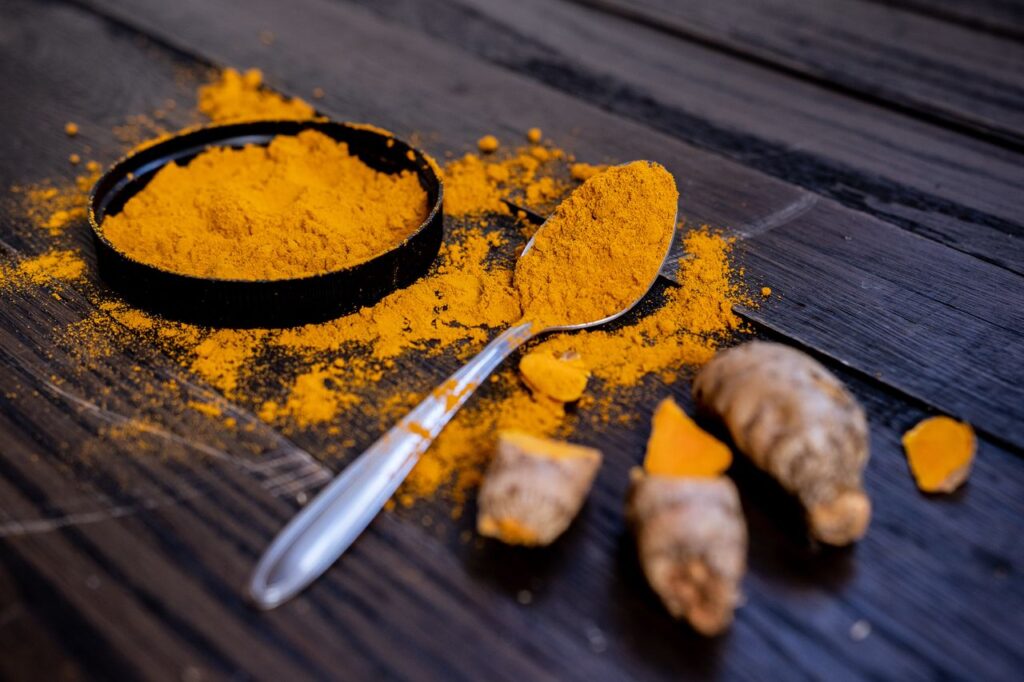 Bright orange ground turmeric in a black dish with a spoon and fresh turmeric root on a dark wooden table