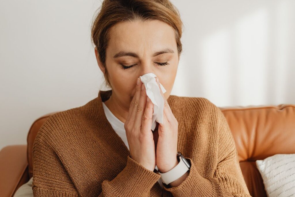 Woman in a brown jumper sitting on a sofa blowing her nose into a tissue with a cold