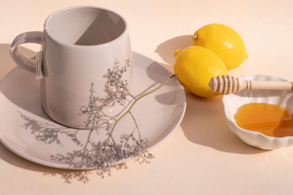 Ceramic mug on a saucer beside two fresh lemons and a small bowl of honey with a wooden dipper