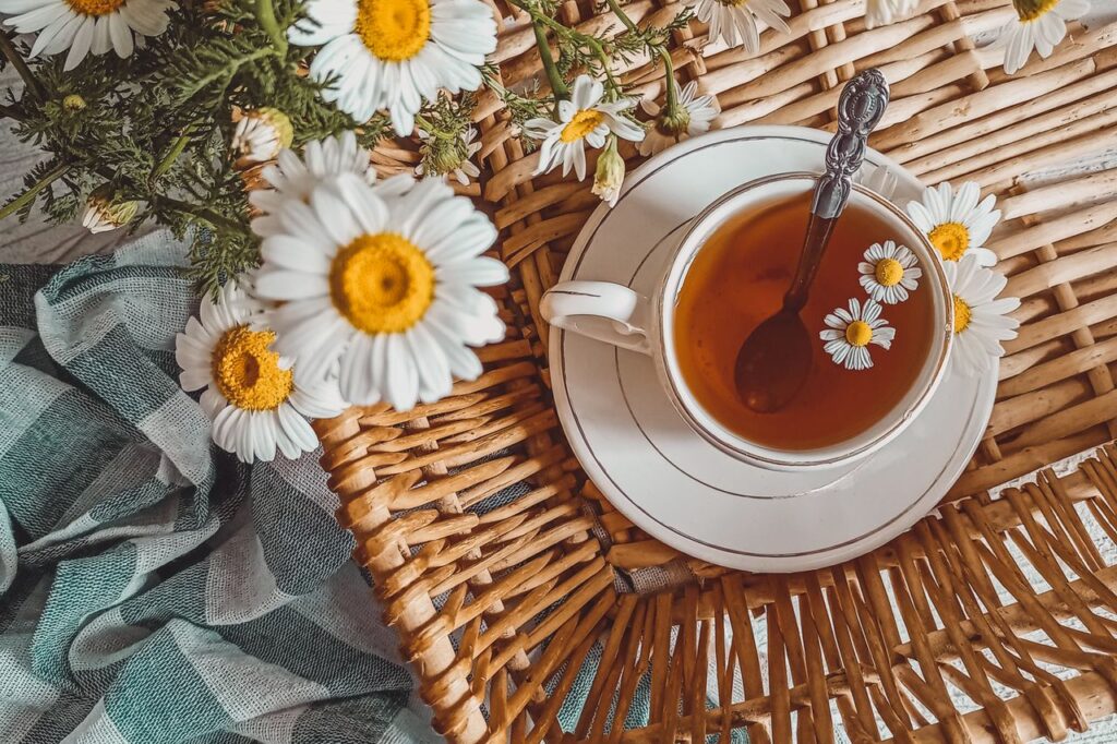 Cup of chamomile tea on a wicker tray surrounded by fresh chamomile flowers with white petals and yellow centres