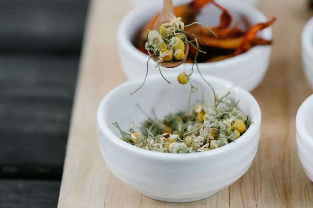 Hand using a small wooden spoon to scoop dried chamomile flowers from a white bowl on a wooden board with other dried herbs