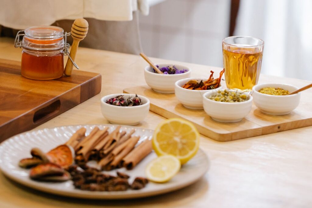 Bowls of dried spices, cinnamon sticks, lemon and a jar of raw honey laid out on a wooden board for making elderberry syrup
