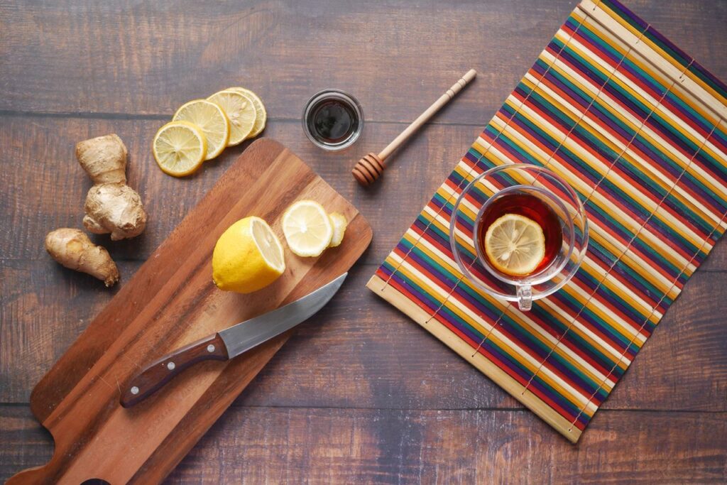 Flat lay of fresh ginger root and lemon slices on a wooden chopping board with a knife, a honey dipper and a cup of ginger tea
