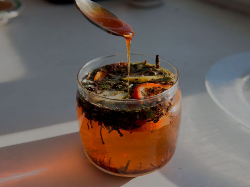 Honey being drizzled from a spoon into a glass of warm citrus tea with lemon and herbs in sunlight