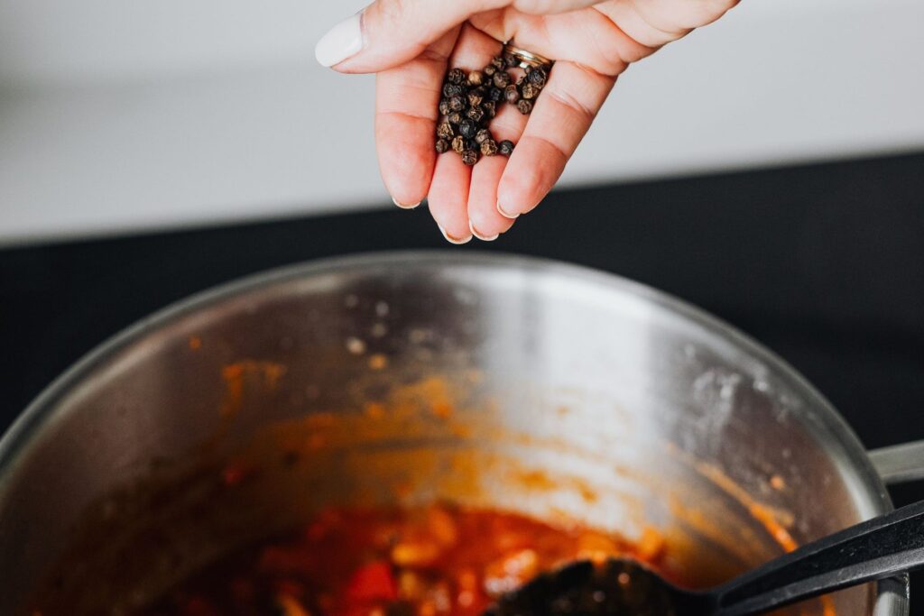 A hand dropping peppercorns into a simmering pot